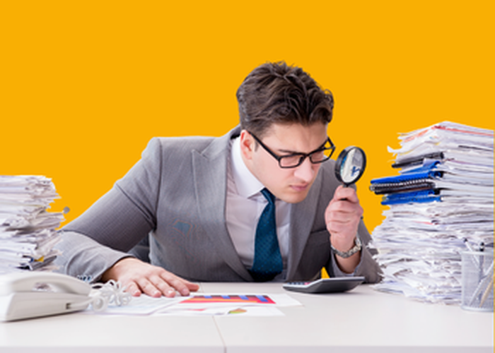 A man buried in paperwork examining documents with a magnifying glass